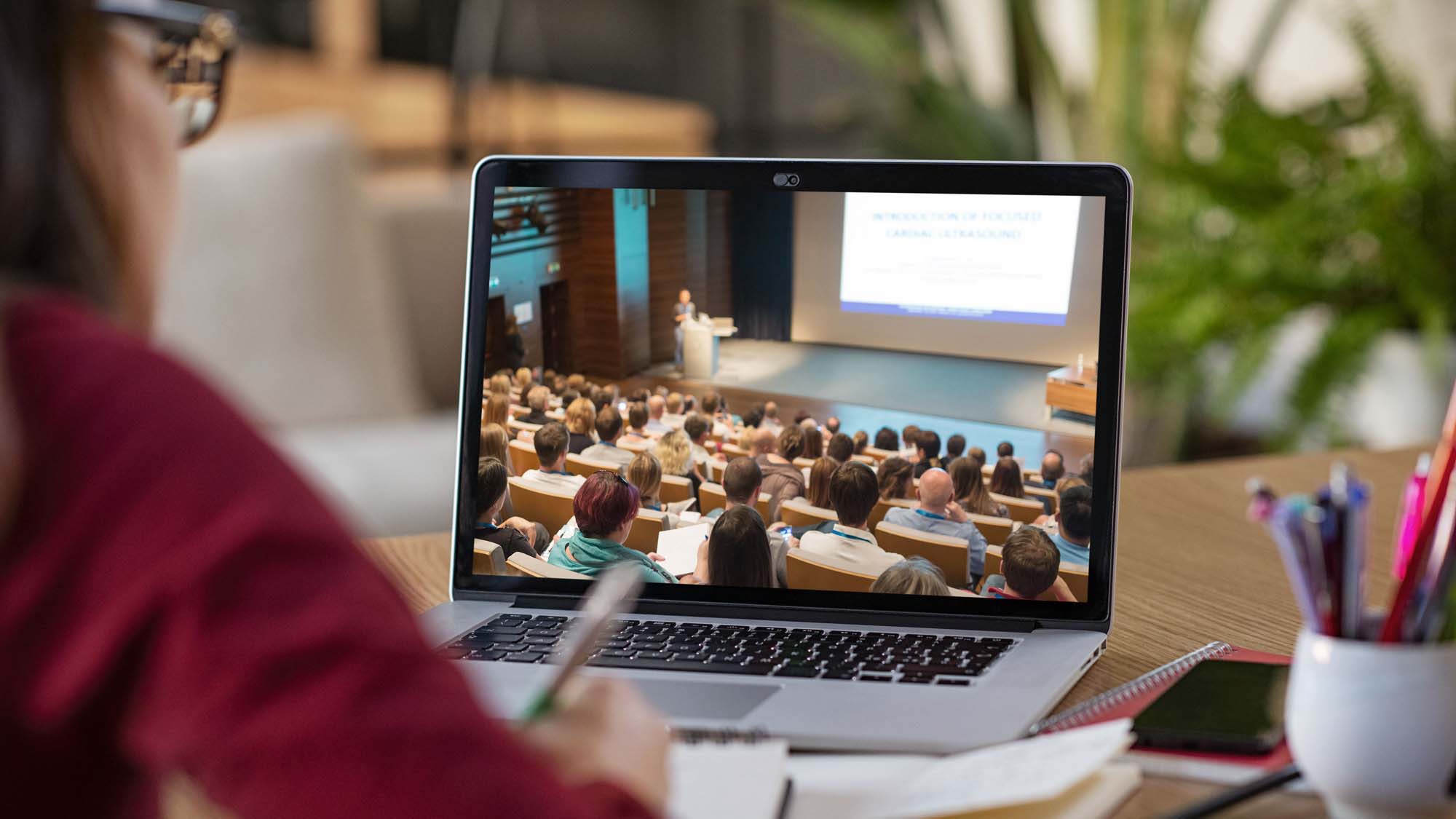 Ringvorlesung-Teaserbild: Person in rotem Pullover schaut auf Laptopbildschirm. Der Bildschirm zeigt einen vollbesetzten Vorlesungssaal.